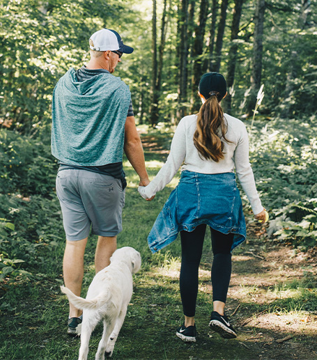 A couple walking in woods holding hands with a white dog walking along side them