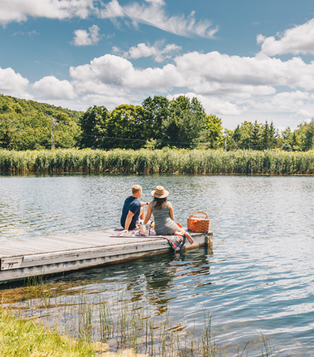 A couple sitting on a small wharf on a lake on clear sunny day with a picnic basket placed beside them
