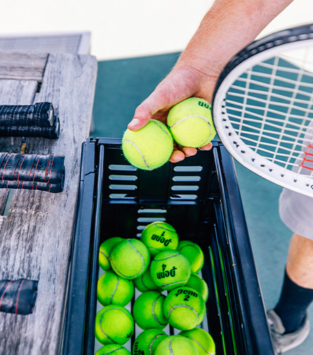 A person with tennis racket picking two tennis balls from a container
