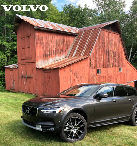 a dark grey volvo SUV on grass with a wooden house in the background