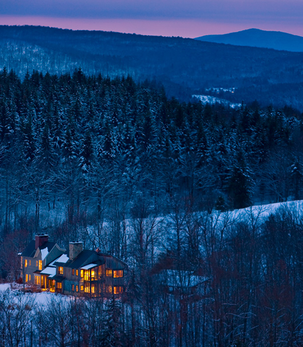 Wooden cottage with lights on at night time in snowy woods with mountains in the background